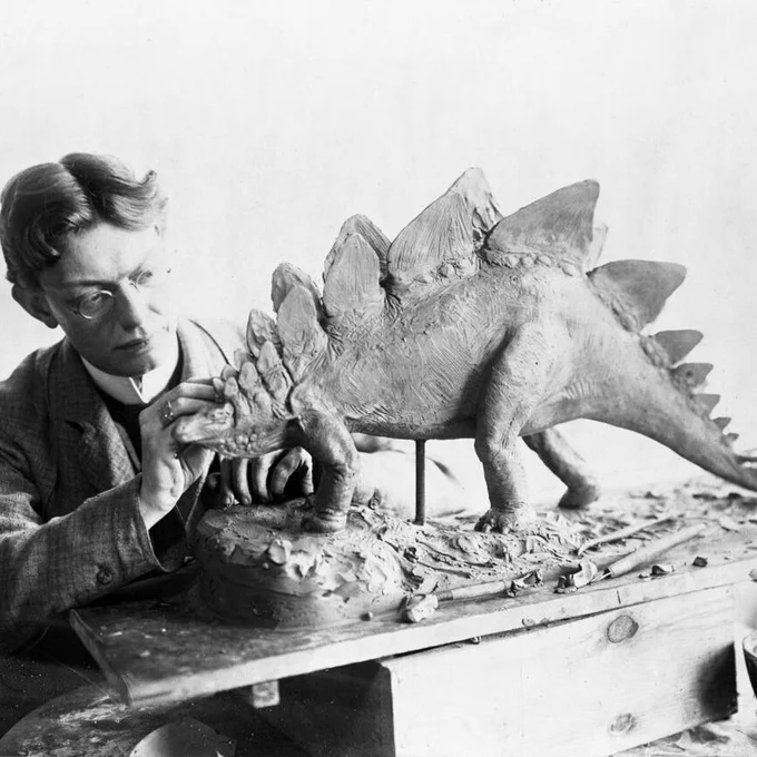 A black and white archival photo of Charles Knight sculpting a model Stegosaurus. The animal has large triangular plates on its back and is depicted dragging its tail, a posture that we know now is inaccurate. 
