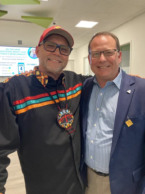 Mike posing and smiling with Clarence, a man wearing a baseball cap, a ribbon shirt and a beaded necklace in modern Indigenous fashion.