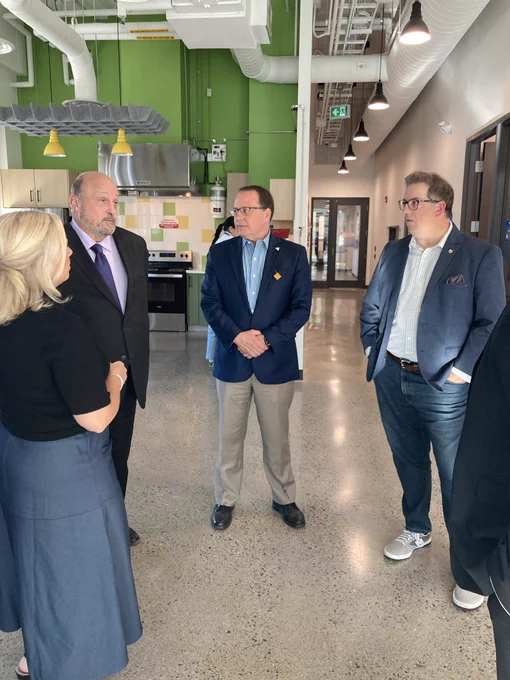 Mike standing with associate minister Michael Tibollo and Mayor Cam Guthrie in a semi circle facing Helen from CMHA in a large space with an industrial kitchen set o in the back.