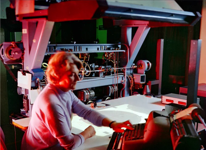 Woman in the foreground typing at a typewriter. Within the background there is a projection of a bubble chamber photograph. This photograph was taken in RHELs High Energy physics laboratory. 