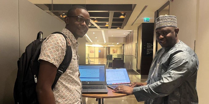 Two individuals standing in a hallway, one wearing a backpack and the other in traditional attire, both smiling and pointing at a laptop screen.