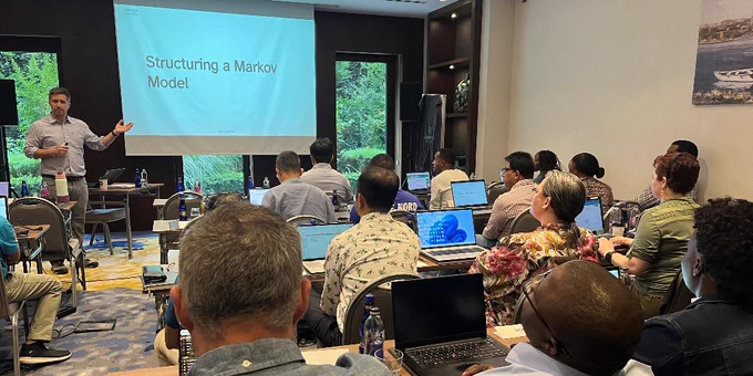 A presenter gestures towards a slide titled "Structuring a Markov Model" at the front of a conference room filled with attentive participants seated at tables with laptops.