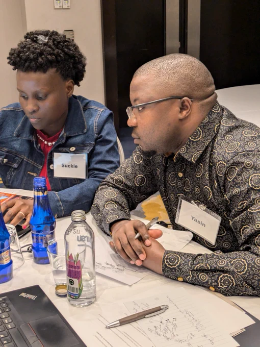 Two individuals named Suckie and Yasin are collaborating at a table filled with papers, laptops, and water bottles, in an indoor setting.