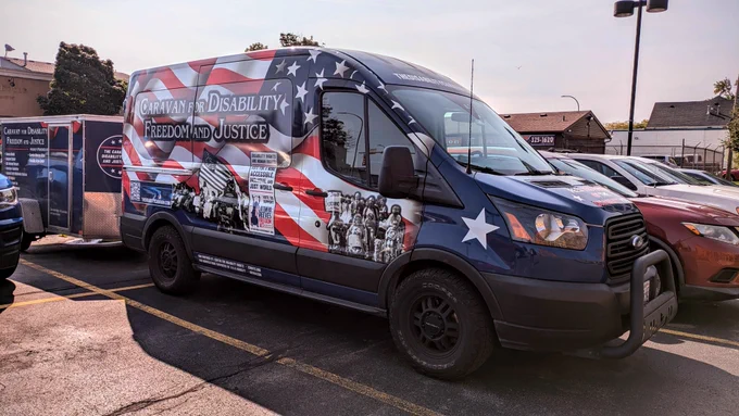 A van parked in the Center for Disability Rights parking lot. The van is painted red, white, and blue with black and white photos of disability activists. The van has the words "Caravan for Disability Freedom and Justice" written on the side.