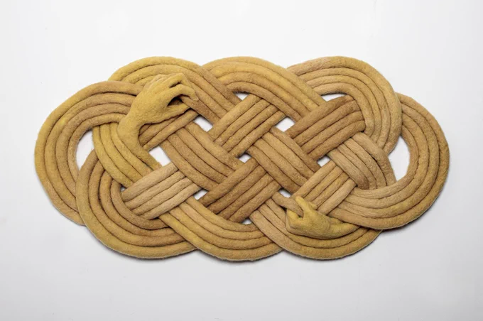 A felted four-strand knotwork oval, with two human hands grasping the stands from within.