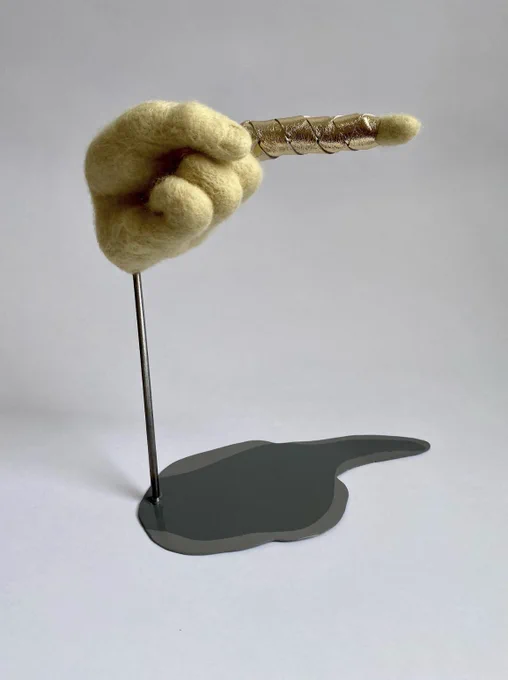 A felted hand with a pointer finger pointed to the side, wrapped in gold leather, akin to a finger trap. The felted hand is elevated above a metal two-tone shadow.