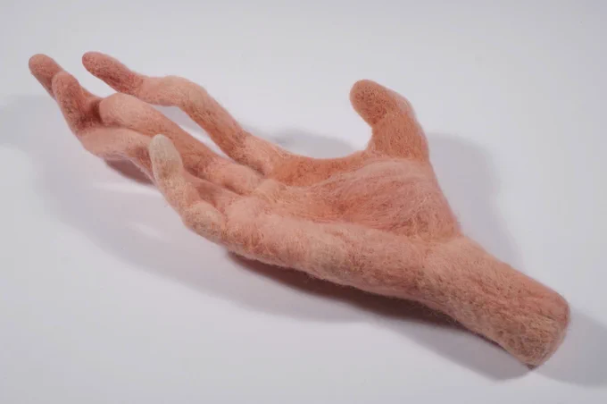 A felted hand raised palm upwards, with three fingers curling and uncannily long.