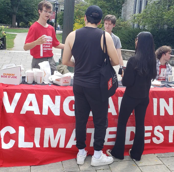 A few people behind a table with a red and white banner speak to a couple of people in front of the table. Photo by Berenice Tompkins