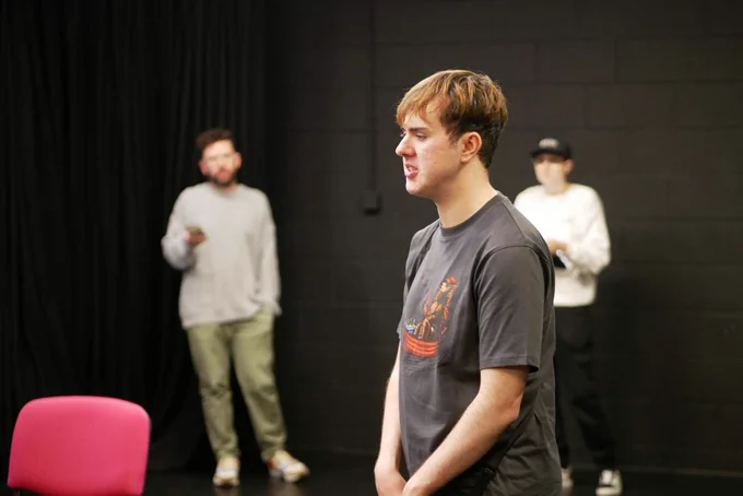A picture of a young man (rehearsing a scene) in the middle of a black rehearsal room looking out to the left (short ginger and brown hair, black t-shirt). Behind him standing near the back wall out of focus, 2 actors look on.