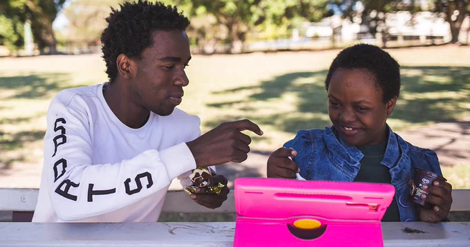 A photograph of a man and a teenager playing with a toy outdoors. Both are  smiling and sitting near a tree. The teen has a pink iPad case propped up in front of them and then are looking directly at the iPad. The man is looking at the teenager. They both seem engaged in a shared fun activity. 