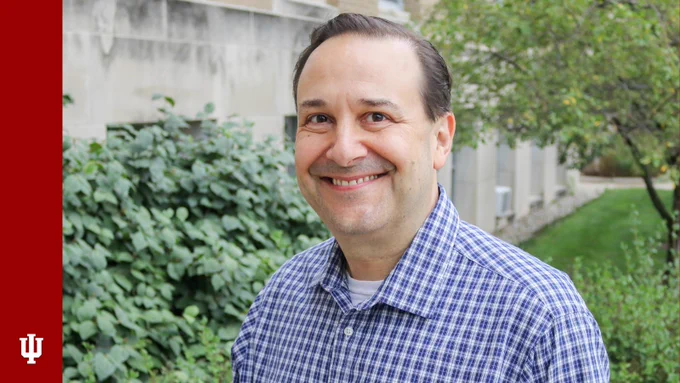 David Haas smiles for his headshot. A limestone building is angled behind him, and trees and bushes frame the photo. A red banner on the left side includes the IU trident.