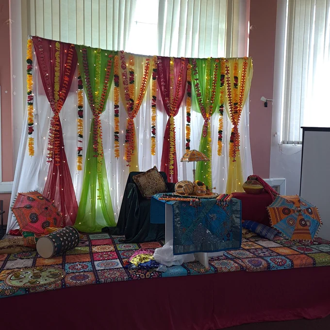 A stage backdrop with colourful hangings and items such as a traditional Asian drum, sequined umbrellas, and multicoloured decorations traditionally seen at South Asian heritage weddings 