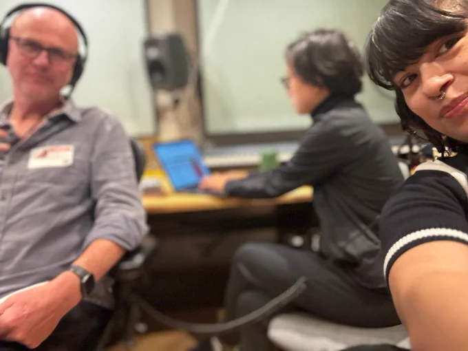 A selfie in a recording studio with one person smiling at the camera and two others working in the background.