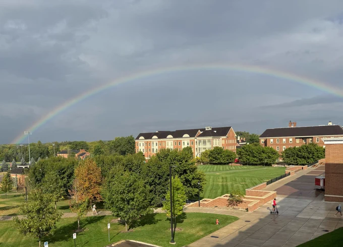 A rainbow stretching over the psychology building of a scenic, tree lined college campus