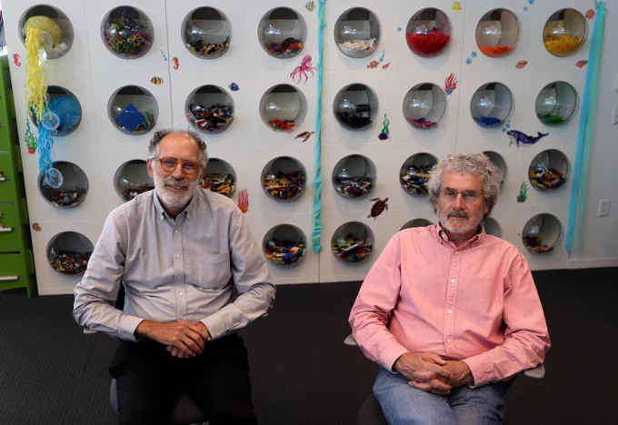 Professors Mitch Resnick (left) and Neil Gershenfeld site in front of a white wall decorated with curved, clear plastic windows containing brightly colored LEGO bricks. Credit: David L. Ryan/Globe Staff
