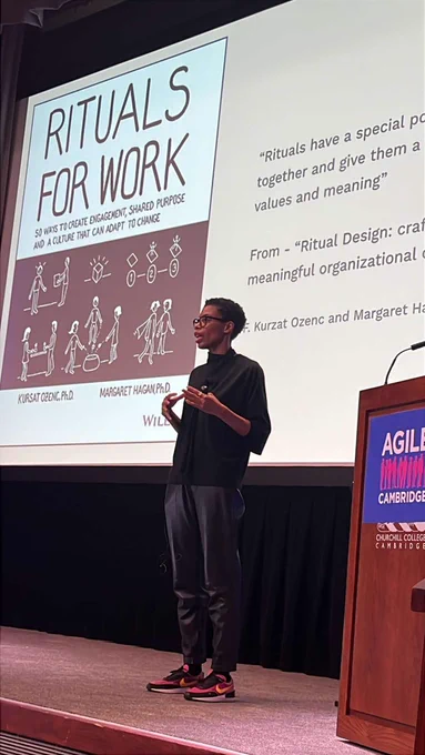 Alison is presenting in front of a large screen displaying a slide titled "Rituals for Work" with various illustrations and text. The event logos for "Agile Cambridge" and "MURAL" are visible at the bottom of the screen.