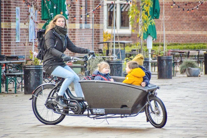 A woman in casual winter attire rides an electric cargo bike with three children sitting in the front bucket on a street in Amsterdam.