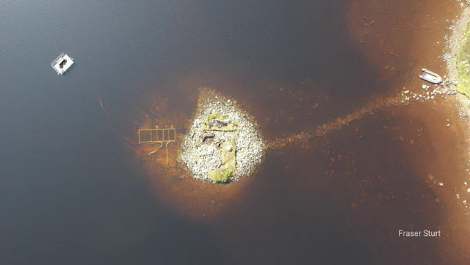 A prehistoric man-made island, or crannog, in Loch Bhorgastail, Scotland. A stone causeway links the crannog to the shore, and remnants of ancient structures are visible on the island. Modern surveying equipment in the water suggests ongoing archaeological research.