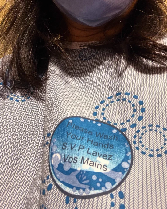 Close up of a blue patterned hospital gown. There is a blue circle that looks like a bubble and it says “Please Wash Your Hands” in English and French. The top of the picture has some black hair and a medical-mask covered chin.