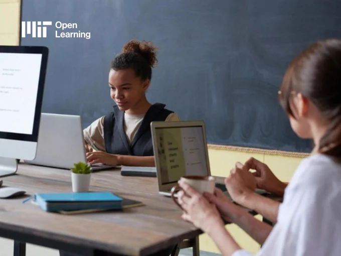 Candid photo of two college students collaborating in a classroom with computers and a chalkboard. The MIT Open Learning logo is displayed in the corner.