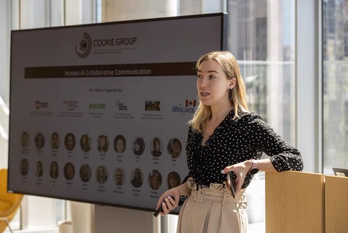 Anastasia Kuzminykh stands at podium holding clicker and speaking in front of screen showing pictures of people in her research group.