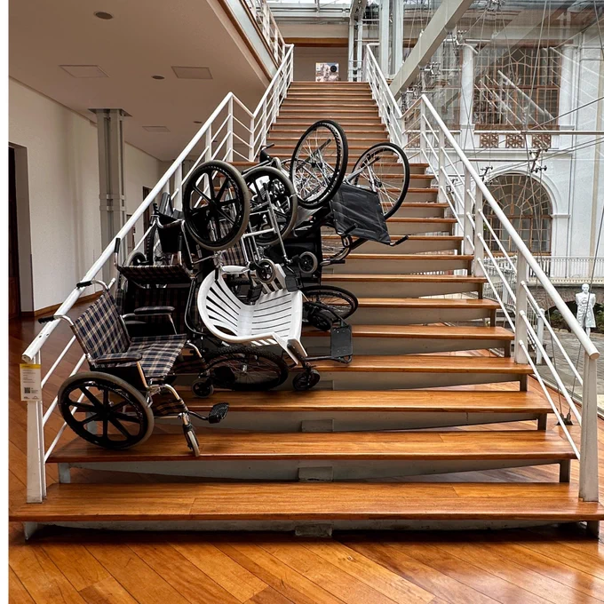 Foto de una escalera donde se ve la obra “In-movilidad”, que consiste en varias sillas y sillas de ruedas apiladas, en representación de la resignificación de los espacios y herramientas de movilidad.