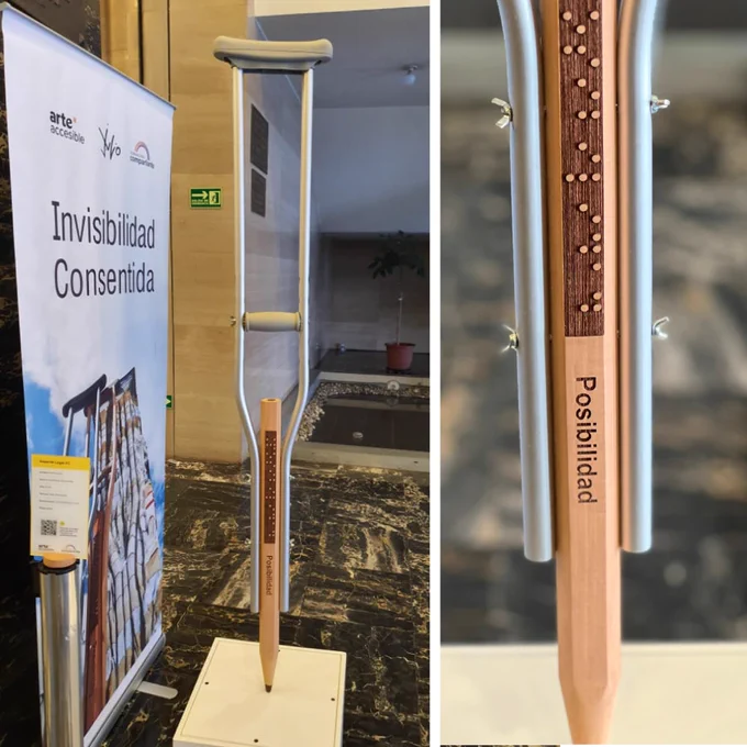 Foto de la obra “Soporte Legal #1”, exhibida en la muestra Invisibilidad Consentida en Quito, Ecuador. La obra presenta una muleta en la que está escrita la palabra “posibilidad”, tanto en letras como en sistema braille. Detrás de la obra hay un banner con el nombre de la muestra, acompañado por el logo del Programa Arte Accesible, la firma del artista Andrés Julio, artista a cargo de la muestra, y el logo de Fundación Comparlante.