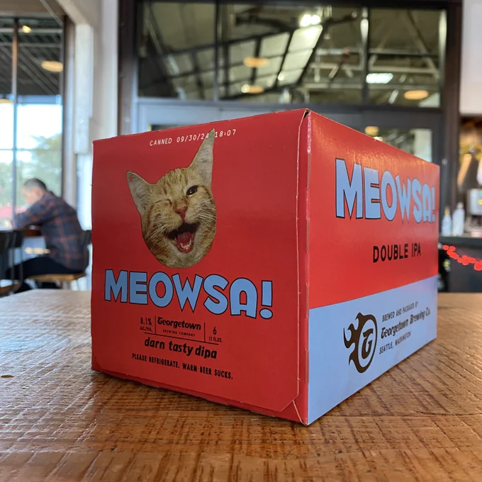 a six pack box sits on a wooden countertop - the box features an orange tabby winking and says Meowsa! Darn tasty DIPA.