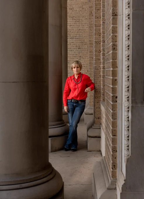Mary Sweitzer standing next to large columns in a red shirt and blue jeans