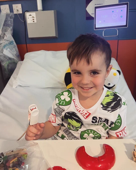 Roman in a hospital bed, smiling with a pulse oximeters, and wearing a colourful Hulk-themed shirt. On the wall behind, a medical monitor displays the time.