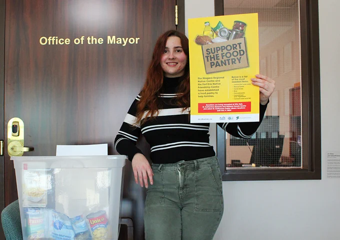 A City staff person in the Mayor's office holds a Support the Food Pantry poster alongside a bin for non-perishable food item donations.