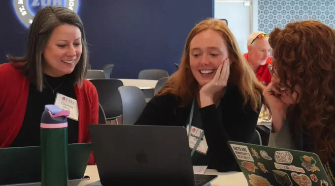 Three participants at a conference, wearing name tags and laughing over a laptop screen. They are seated at a table with various personal items visible, including a water bottle and a laptop covered in stickers.