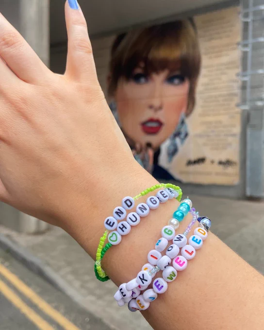 A close-up photo of a person's hand, who is wearing a stack of friendship bracelets. The top two bracelets are green and say 'End hunger'. In the background is a mural of Taylor Swift painted on a wall at Wembley.