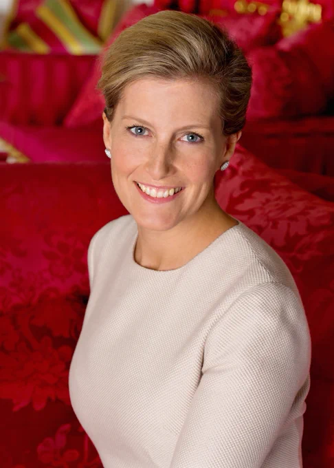 Her Royal Highness The Duchess of Edinburgh sitting on a red sofa with floral pattern. She’s looking directly at the camera and smiling.