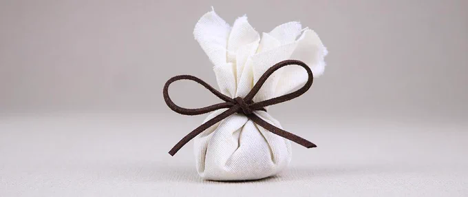 This image shows a small white fabric pouch tied with a brown string into a neat bow. The fabric appears soft and has a slightly rustic, handmade feel, with its edges gently fraying. The overall look gives a sense of simplicity, elegance, and natural charm.