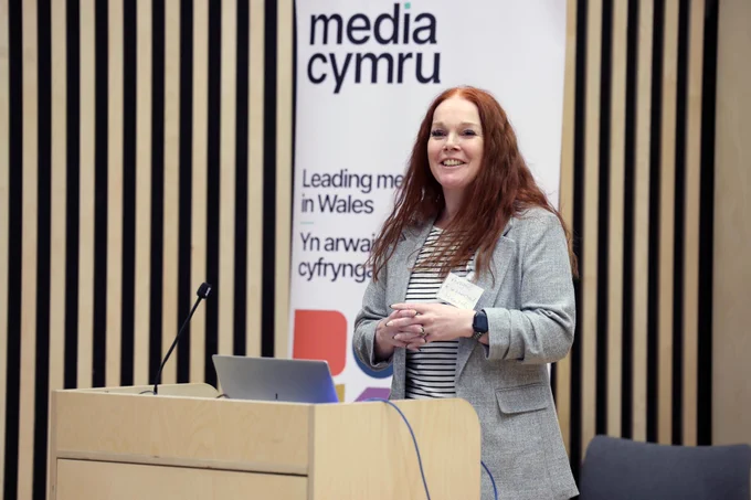 a woman with red hair standing behind a podium presenting to an audience, there is a banner behind her which reads media cymru