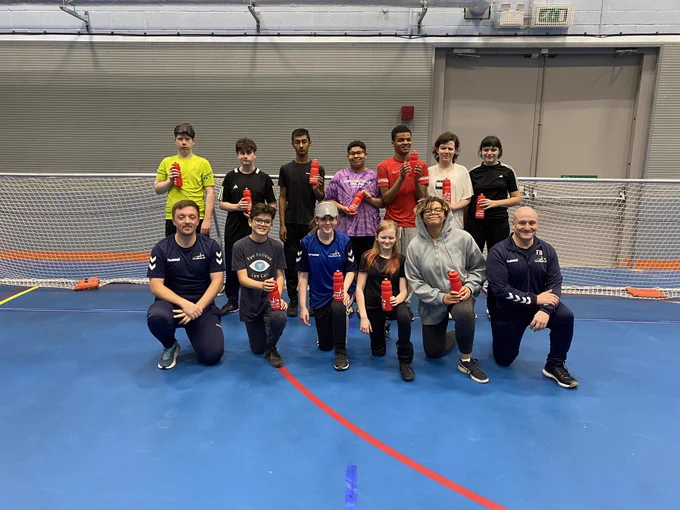 Group photo of all attendees smiling with players and coaches in the photo, across two rows. Players are each holding a water bottle they received for attending the day. Everyone’s in front of a goalball goal.