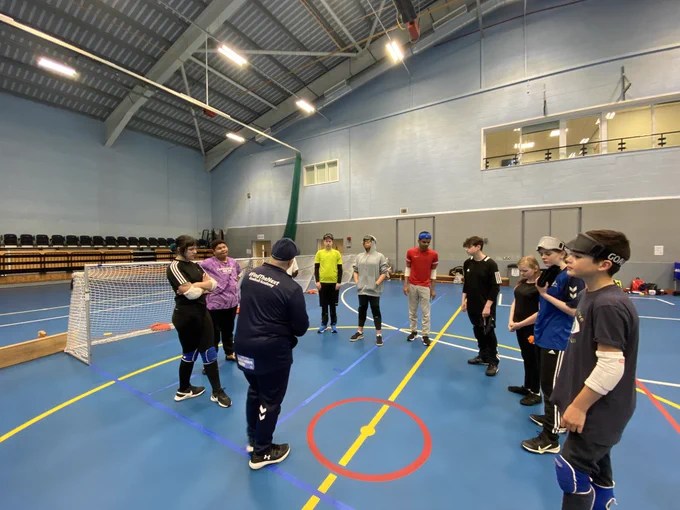 Coach Tommy speaking to the group attending our RNC Academy Aspire Day, with 11 players all ready to play goalball stood in a circle in a sports hall. Tommy is outlining the next part of the session.