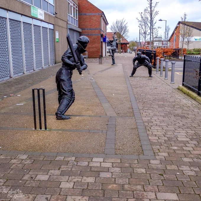 Three metallic statues of cricketers are acting out the 1932-33 Bodyline Ashes series, on an empty brick-laden town square in front of a shuttered-up library. The view is from behind the batter, first slip, maybe.