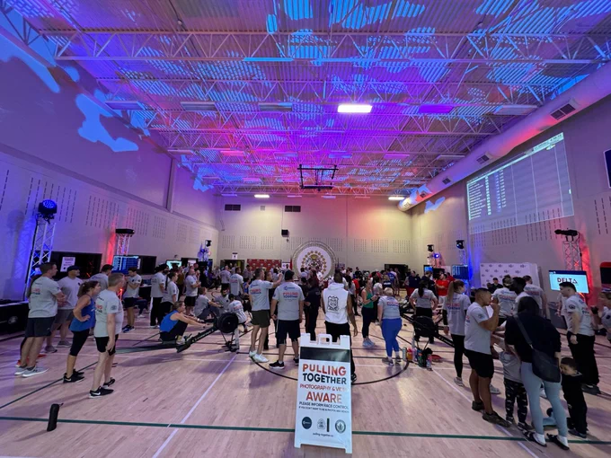 A large group of people inside a gym participate in Pulling Together, with some people rowing and others waiting their turn. 
