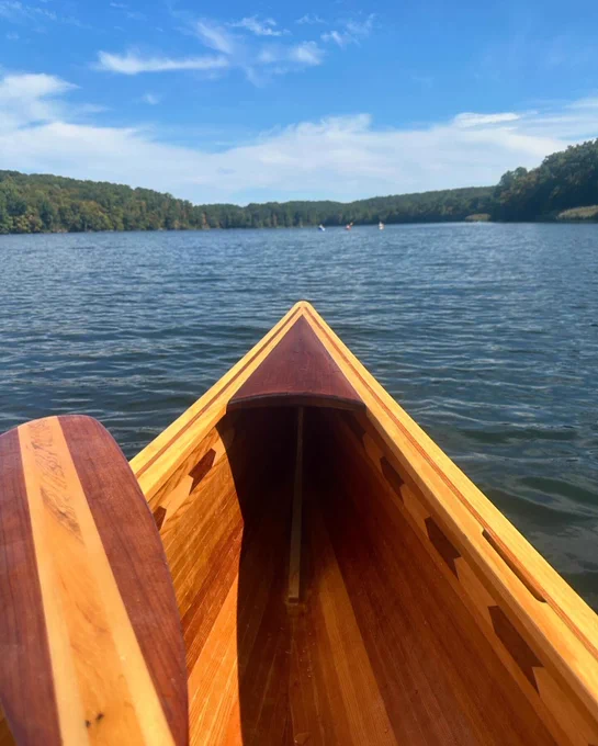 The bow of a cedar strip canoe takes center stage.  The blade of one oar is visible resting on the gunwale. The water is rippled by the wind. In the distance are green trees all around the shoreline. Way in the distance you can just make out three kayaks.