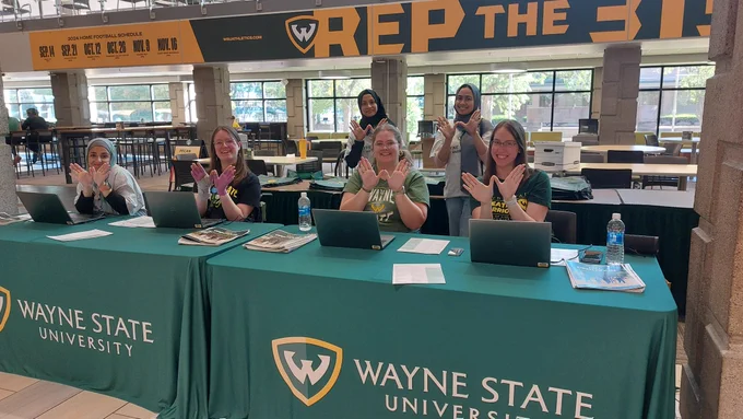 A group of Wayne State students sitting at a table that says Wayne State University and holding up the W sign