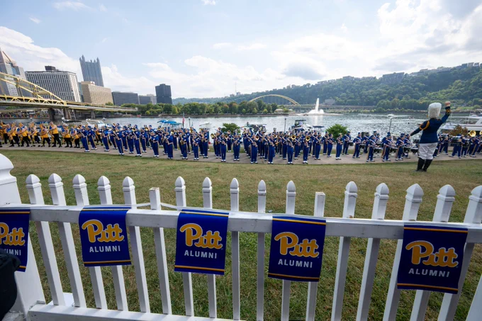 Members of the University of Pittsburgh marching band and alumni gather outdoors for an event, with the Pittsburgh skyline in the background.
