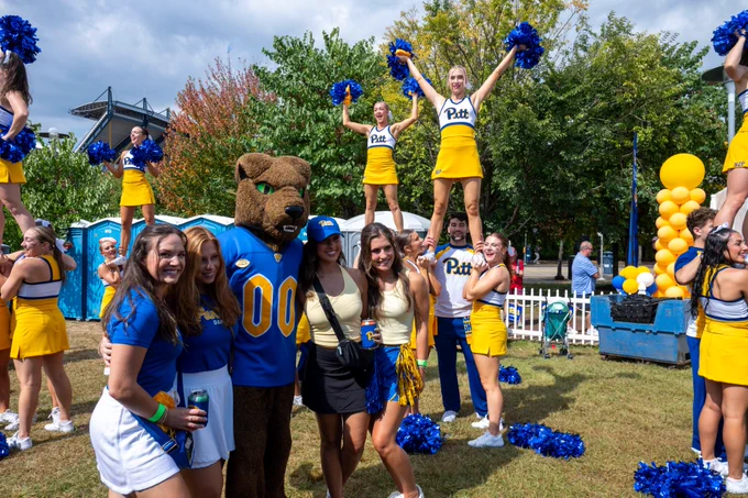 Group of University of Pittsburgh cheerleaders and fans posing with the mascot at an outdoor event, surrounded by blue and gold decorations.