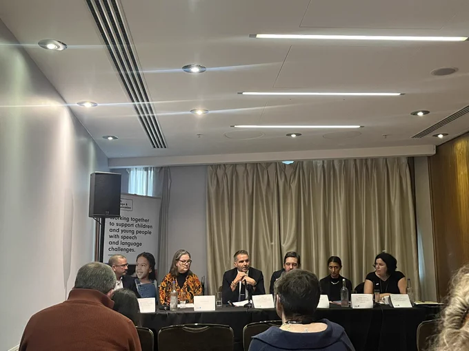 Speech and Language UK panel. 6 people sit at a table in a room of people. Green curtain behind them.