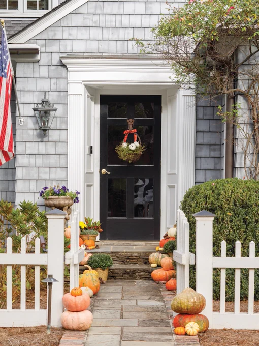 Alt text: The entryway to a gray-and-white shingle house with a black door, framed by a white picket fence, an American flag, a basket hung on the door holding pumpkins, and a collection of pumpkins and gourds lining the sidewalk and front steps. 