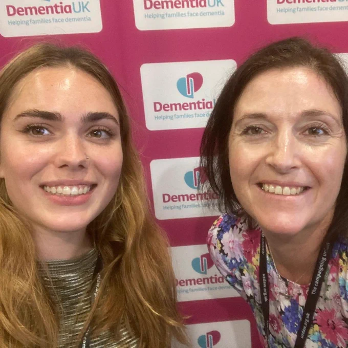Two individuals smiling, standing in front of a banner with the Dementia UK logo and the phrase “Helping families face dementia.”