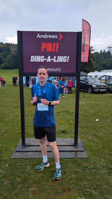 Matt wearing a blue top and black shorts with turquoise green running shoes standing in front a purple sign with a black border which says "PB!! DING-A-LING! Run Reigate" 