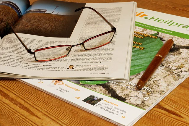 A wooden table with a magazine open with a pair of glasses and a pen on top.