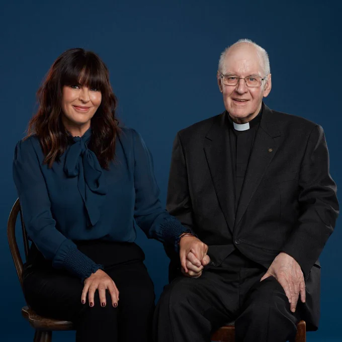 Anna Richardson and her father, Jim, sit on some chairs in front of a blank teal wall. They hold hands and smile for the camera. Anna has long, dark, wavy hair with a blunt fringe. She wears a teal top and black trousers. Jim is an older man with short white hair. He wears glasses and the black suit, and white collar of a vicar.